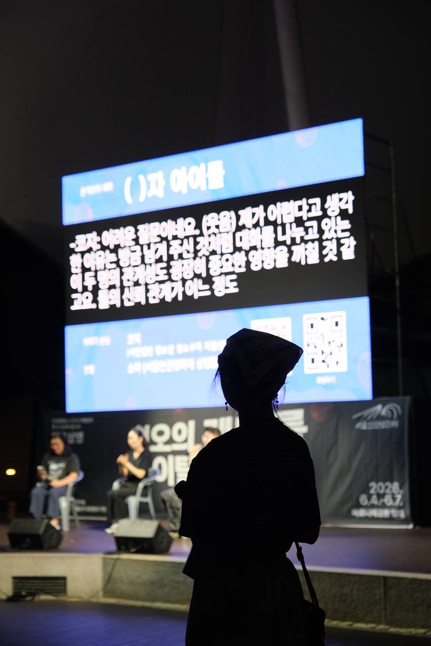 사진. 영화 "()자 아이들" 관객과의 대화가 진행 중인 마로니에공원 야외무대. 무대와 스크린을 보는 이의 뒷모습 실루엣.