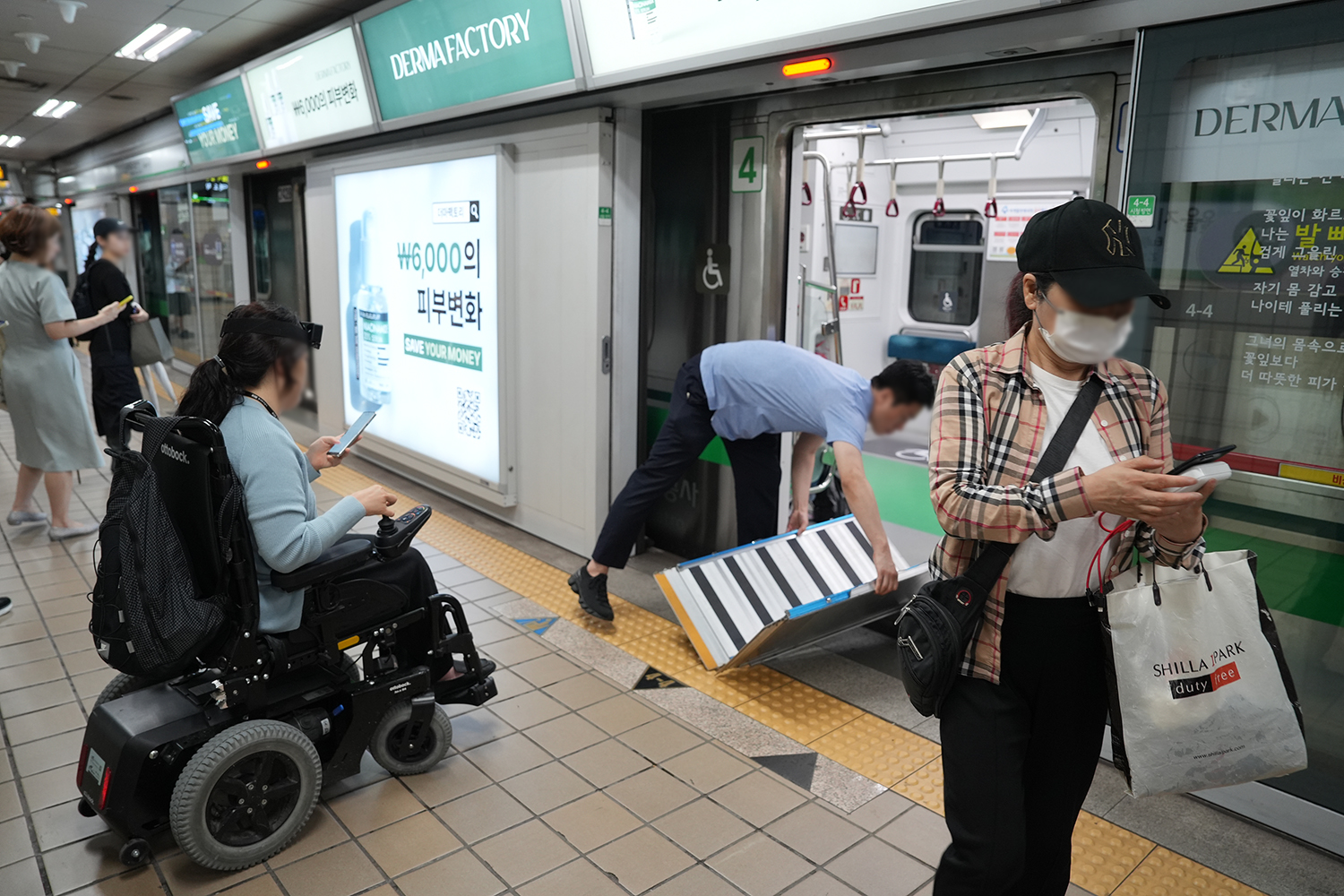 지하철 승강장에서 전동휠체어 이용자가 지하철에 탑승하려고 하는 장면. 전동차 문이 열려있고 역무원이 접이식 발판을 설치하고 있다. 스크린도어에 광고들이 부착돼있고 주변에 보행자가 있다.