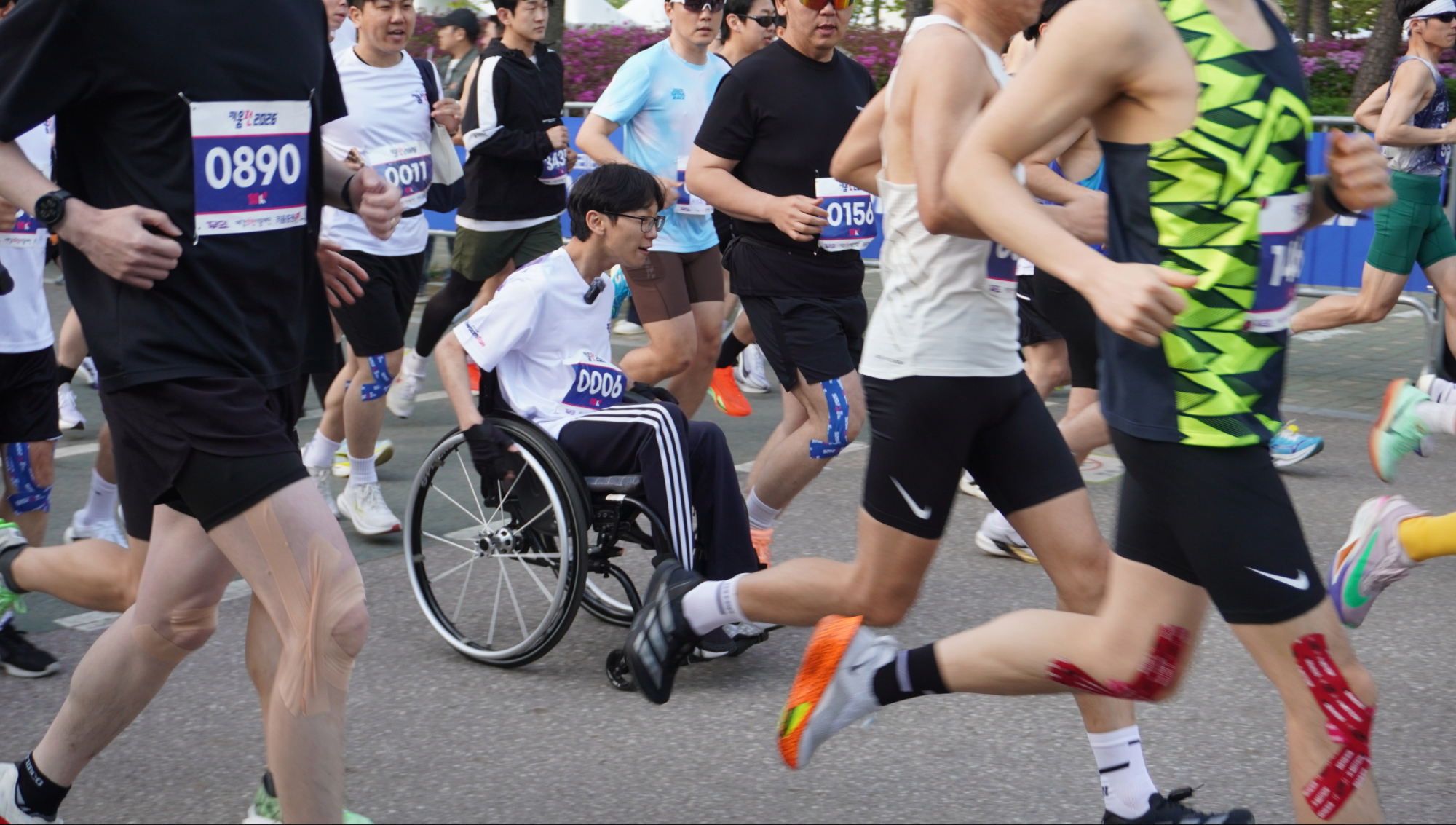 가운데 수동휠체어를 이용하는 흰색 상의, 검정색 하의를 입고 안경을 낀 러너가 있고 그 주위에 형형색색의 다양한 옷을 입은 러너들이 힘차게 달리고 있다.