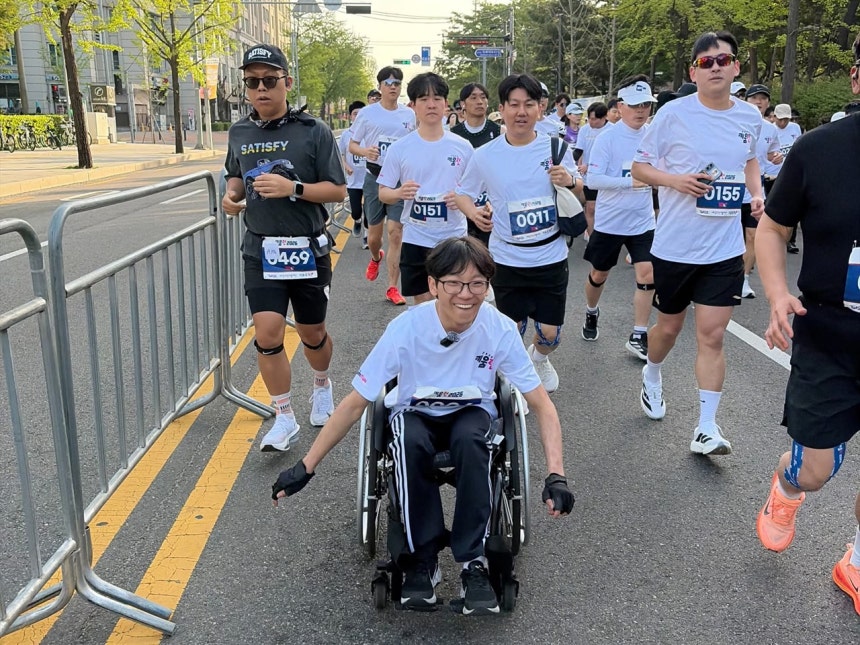 수동휠체어를 이용하고 흰색 상의에 검정색 하의를 입고 안경을 낀 러너가 가운데 있고 그 주변에 다양한 옷을 입은 러너들이 달리는 사진.