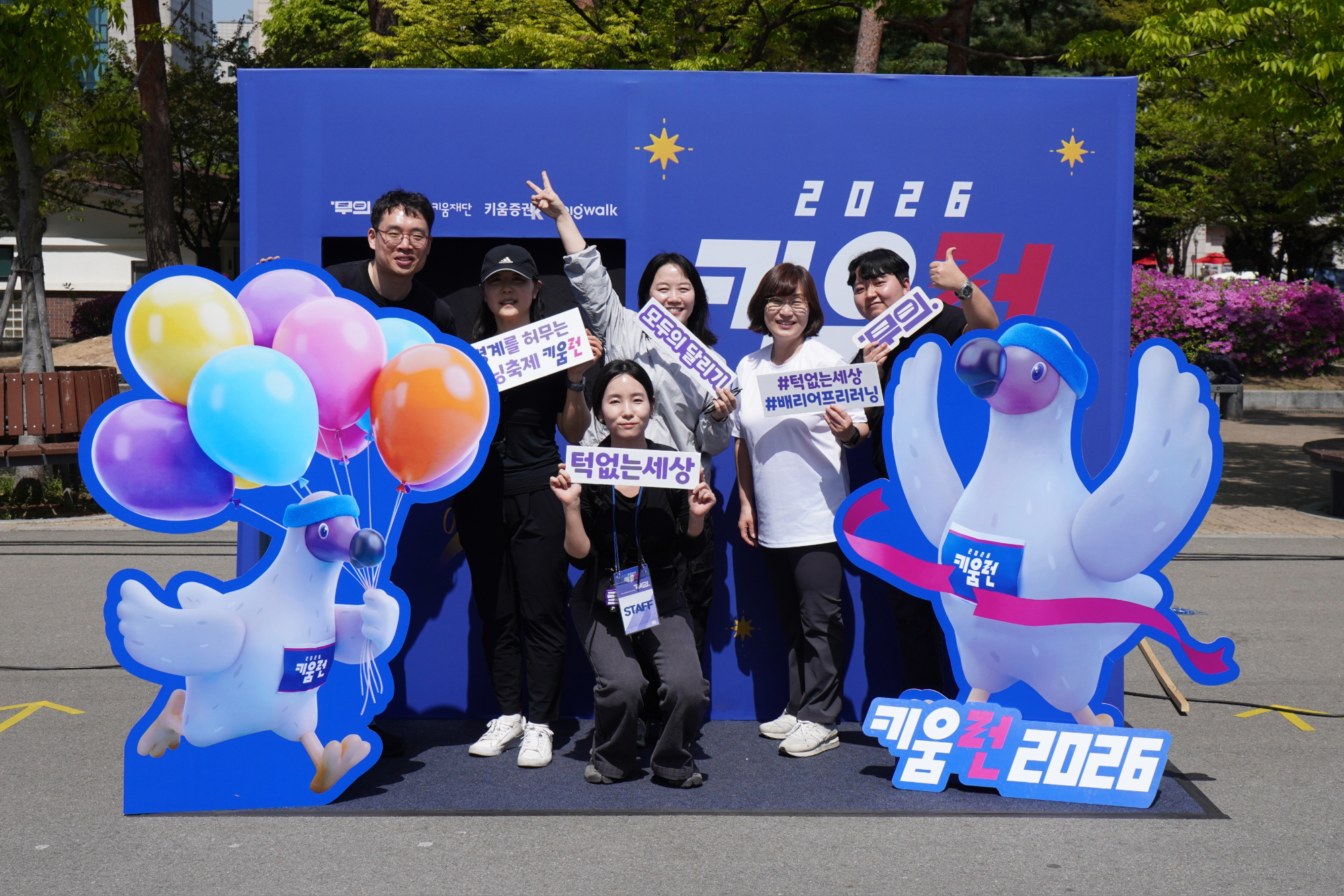 파란색 2026 키움런 배경 앞에 무의 구성원들이 다양한 글씨가 적힌 피켓들을 들고 서있다. 양 옆으로 풍선을 든 도도새와 양 날개를 번쩍 든 도도새가 있다.