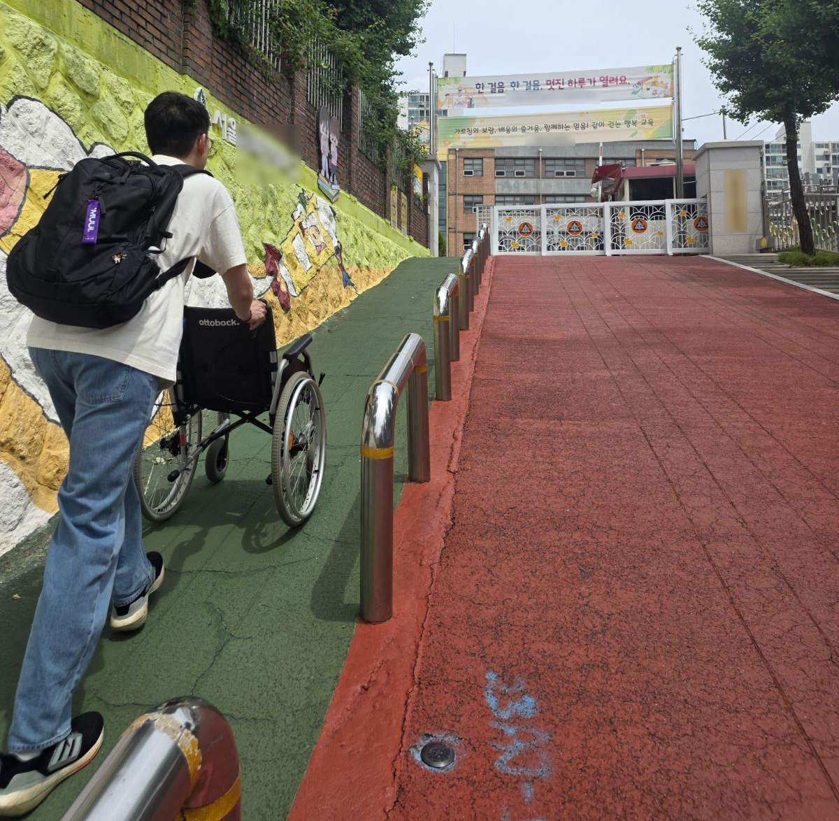 경사가 심한 학교 정문 앞 길을 검정색 백팩을 맨 성인 남성이 빈 수동 휠체어를 밀며 올라가고있다.