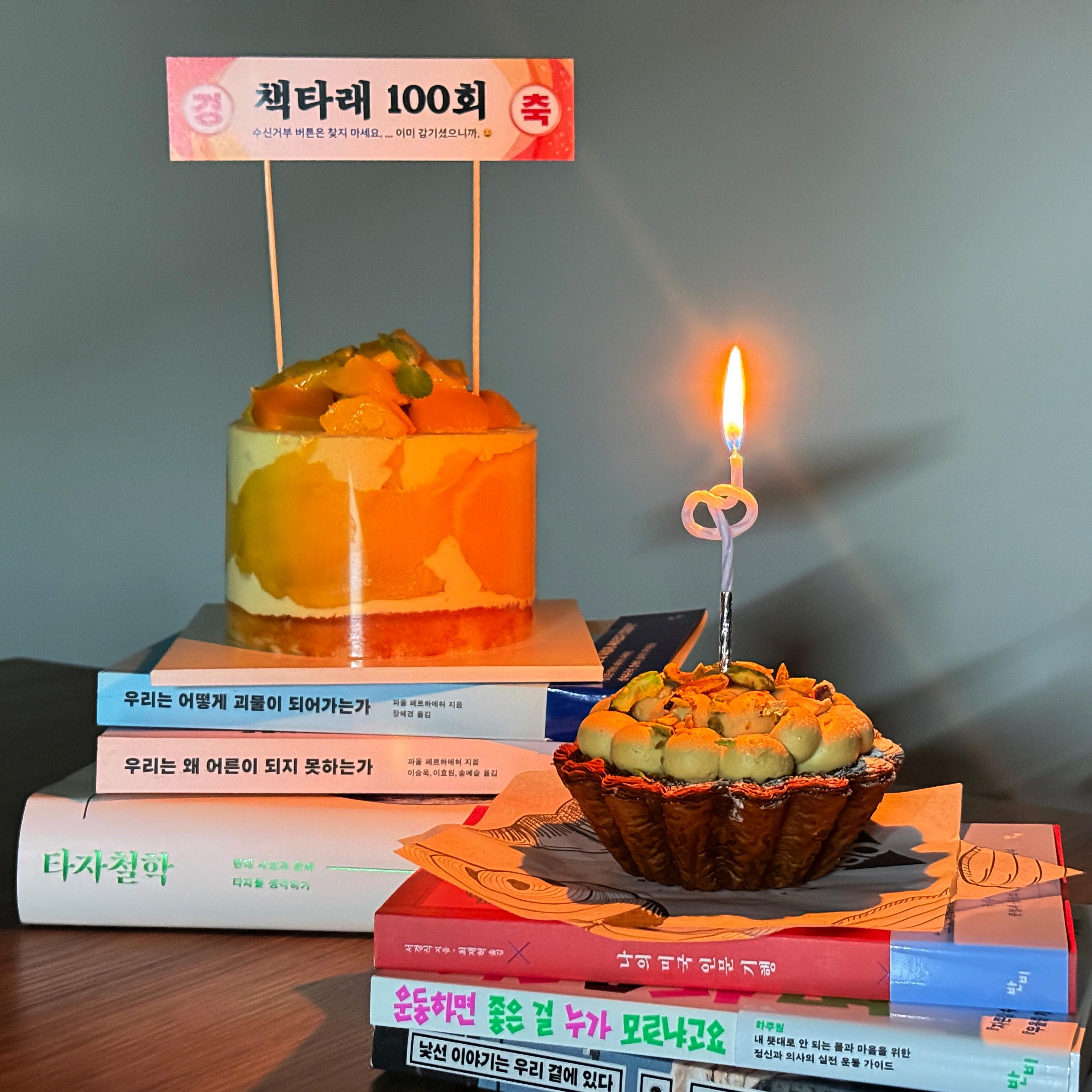 반비 책 위에 놓여 있는 망고케이크와 피스타치오 플랑(타르트) . 망고케이크에는 책타래 100회를 축하하는 플래카드 디자인의 장식이 꽂혀 있다. 내용은 "경) 책타래 100회 (축/수신거부 버튼은 찾지 마세요/...이미 감기셨으니까. : D"